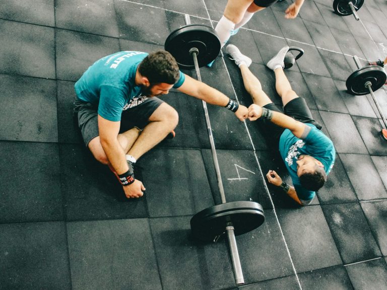 Two men in workout gear, one assisting the other on the gym floor near a barbell.